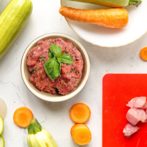 Photo of carrots and other vegetables surrounding a white dog bowl with Drool Pet Co. Beef best dog food with basil as garnish